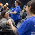 Two women, one standing the other in a wheelchair, hold hands and smile while dancing. Other dancers are in the background.