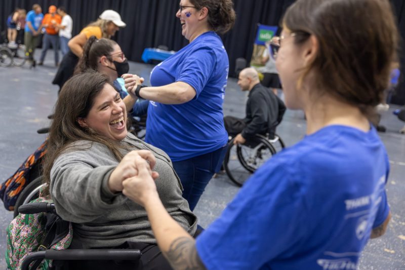 Two women, one standing the other in a wheelchair, hold hands and smile while dancing. Other dancers are in the background.