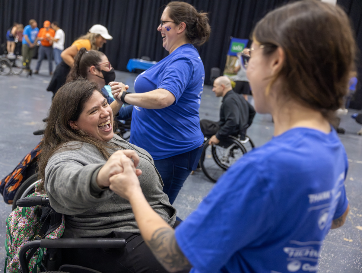 Two women, one standing the other in a wheelchair, hold hands and smile while dancing. Other dancers are in the background.