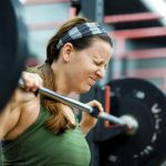 A woman closes her eyes as she lifts a heavy weight off of a rack at a gym.