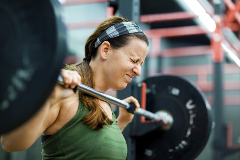 A woman closes her eyes as she lifts a heavy weight off of a rack at a gym.