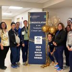 Thirteen members of the Neuroscience Critical Care team at Milton S. Hershey Medical Center smile as they stand around a pull-up banner that reads “Congratulations to our Neuroscience Critical Care – 2024 Gold Beacon Award Winners.”