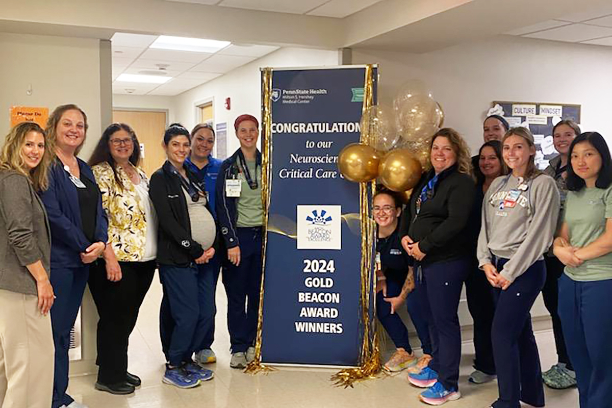 Thirteen members of the Neuroscience Critical Care team at Milton S. Hershey Medical Center smile as they stand around a pull-up banner that reads “Congratulations to our Neuroscience Critical Care – 2024 Gold Beacon Award Winners.”