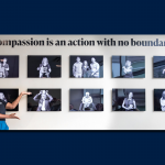 A Woman stands far to one side with her back to a display of photographs on a wall as she gestures for people to look at the photos behind her.