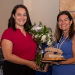 Featured Image Two ladies pose side-by-side, smiling. One holds a vase of flowers, the other hands her a Penn State Nittany Lion trophy.