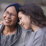 A younger woman embraces a senior woman with her right arm.