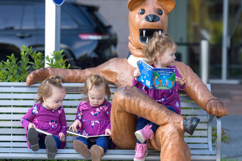 Three young girls, all dressed in identical outfits, sit on a bench with a Nittany Lion statue. One of the girls sits in the Lion’s lap, the other two are just to his side. A vehicle and a building are in the background, slightly out of focus.