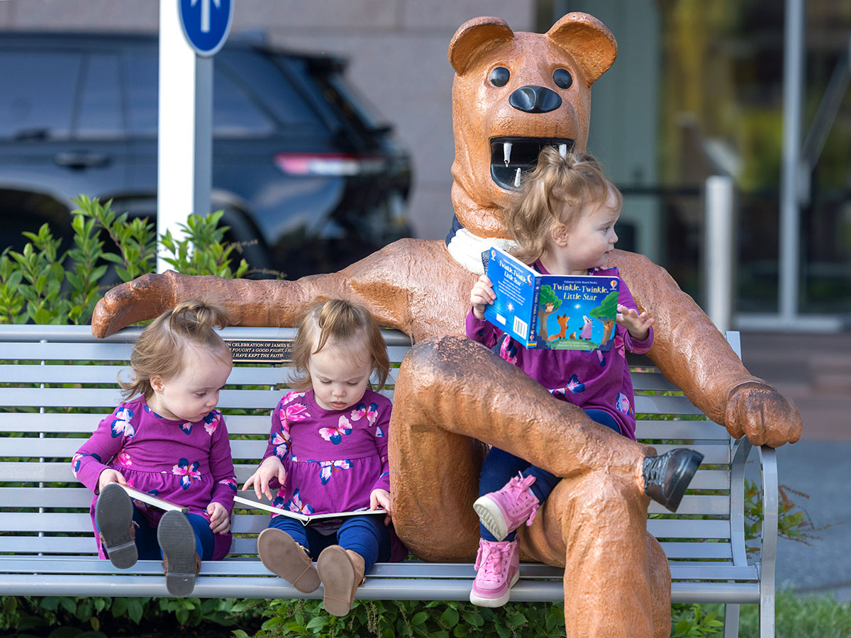 Three young girls, all dressed in identical outfits, sit on a bench with a Nittany Lion statue. One of the girls sits in the Lion’s lap, the other two are just to his side. A vehicle and a building are in the background, slightly out of focus.