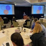 Dr. Fabio Cominelli stands at a podium, flanked on both sides by projector screens, in a room full of people seated at tables.