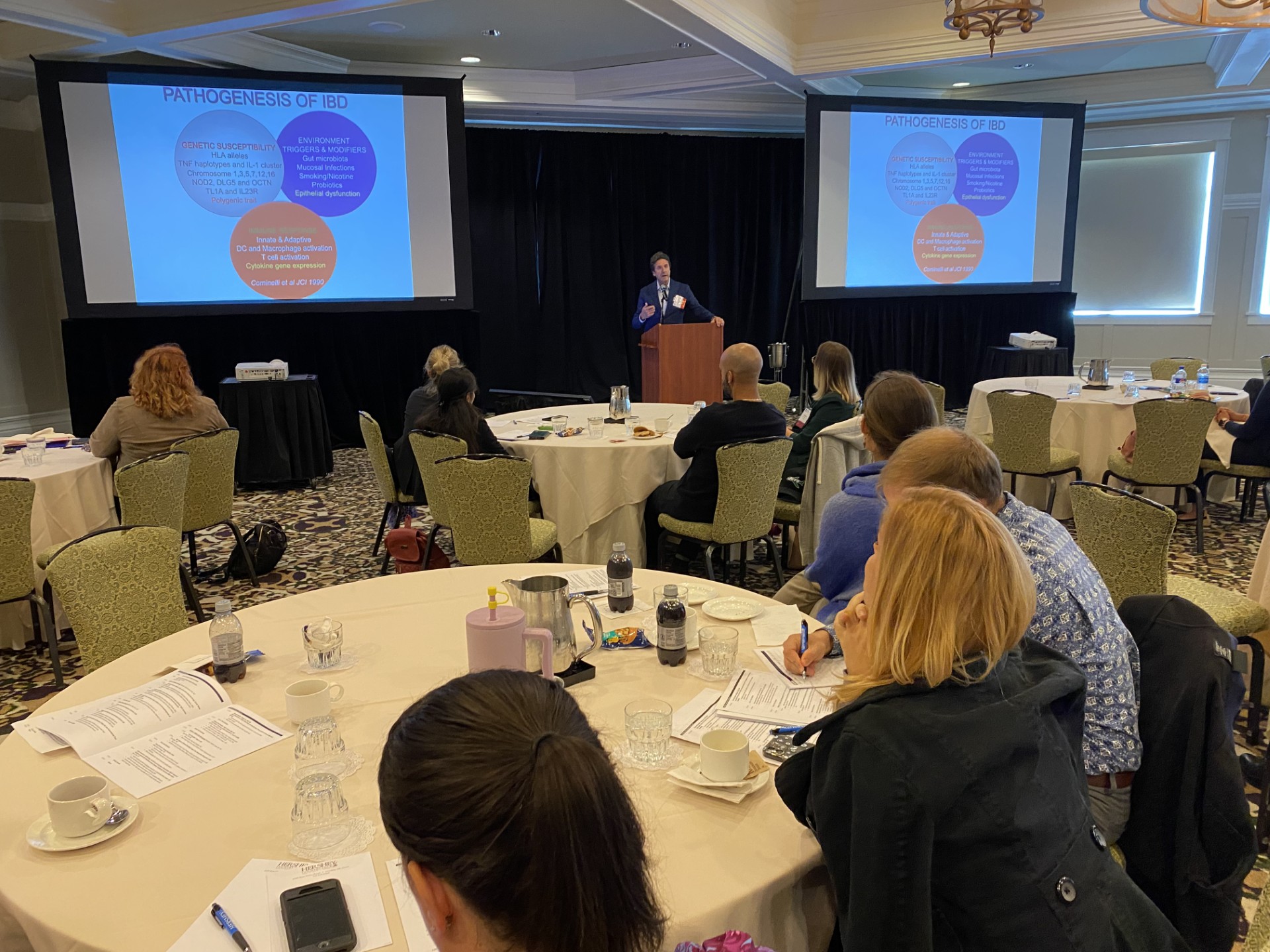 Dr. Fabio Cominelli stands at a podium, flanked on both sides by projector screens, in a room full of people seated at tables.