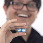 Joseph Romero poses with a pair of specially designed dice that he’s holding near the camera. Joseph is in the near background, slightly out of focus, smiling.