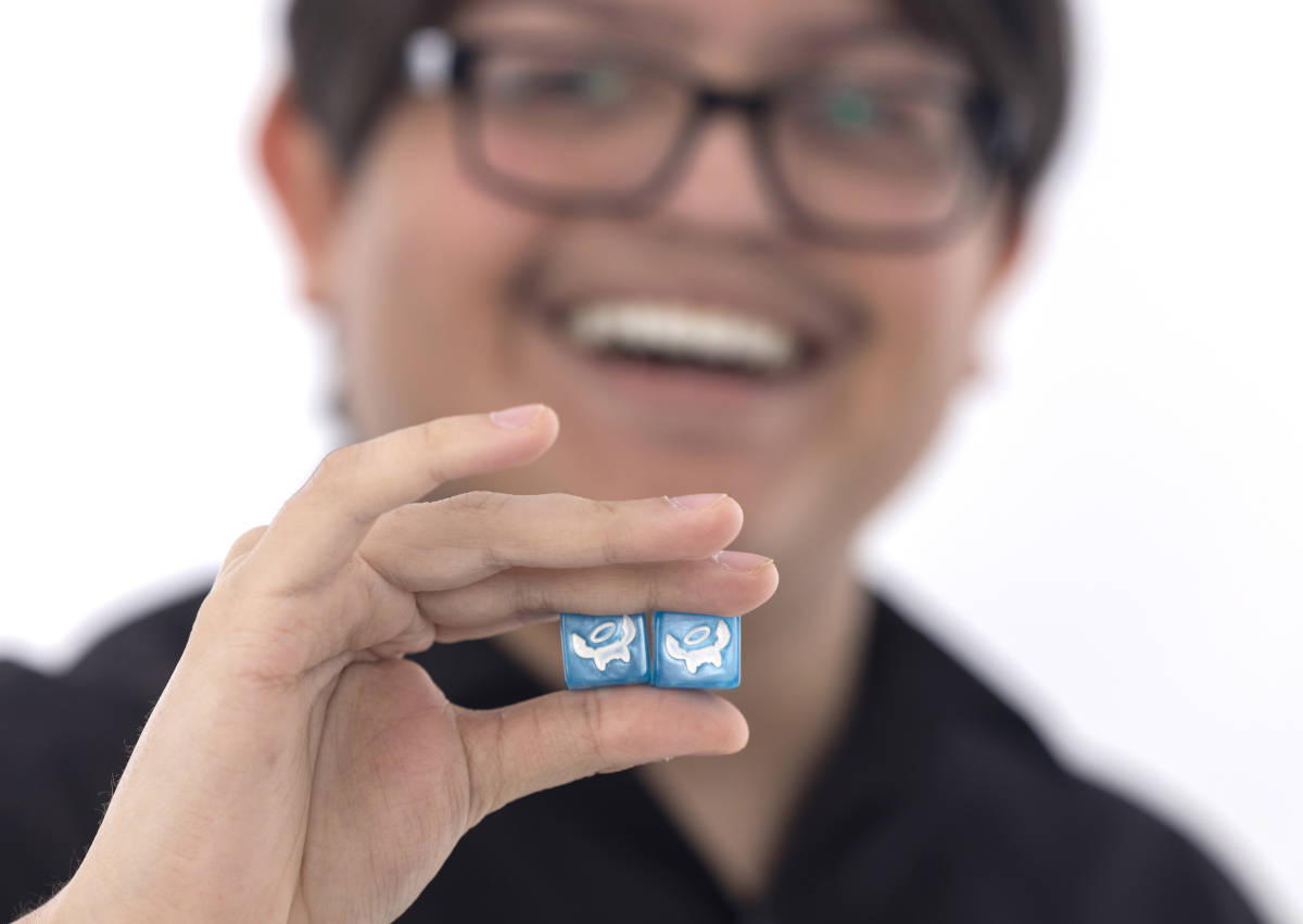 Joseph Romero poses with a pair of specially designed dice that he’s holding near the camera. Joseph is in the near background, slightly out of focus, smiling.