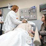 Two nurses tend to a mannequin posing as a patient while a woman sits at the bedside, looking on.