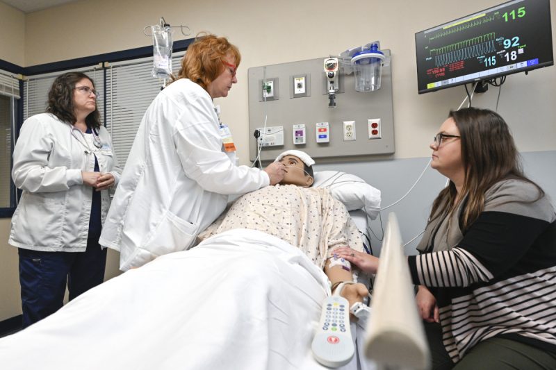 Two nurses tend to a mannequin posing as a patient while a woman sits at the bedside, looking on.