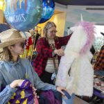 A mother and her teenage son peruse costumes and other items laying on tables. The son, seated in a wheelchair, holds balloons and other items.