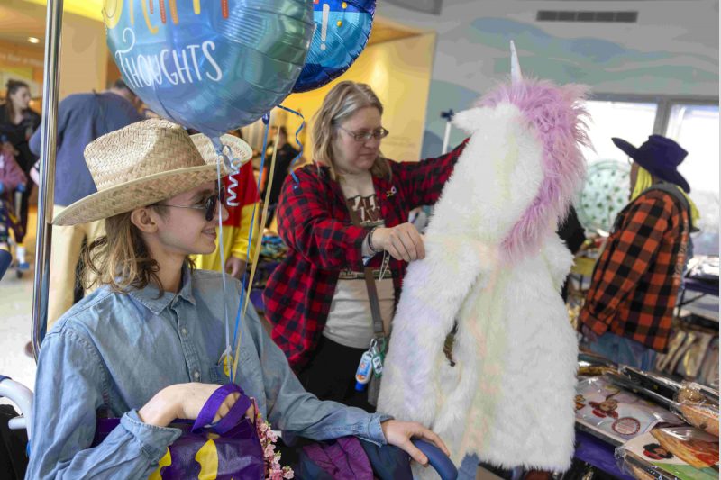 A mother and her teenage son peruse costumes and other items laying on tables. The son, seated in a wheelchair, holds balloons and other items.