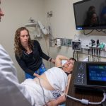 A physician assistant practices an ultrasound on a patient laying in a bed. A man in a white coat stands to the side looking at a medical device screen.