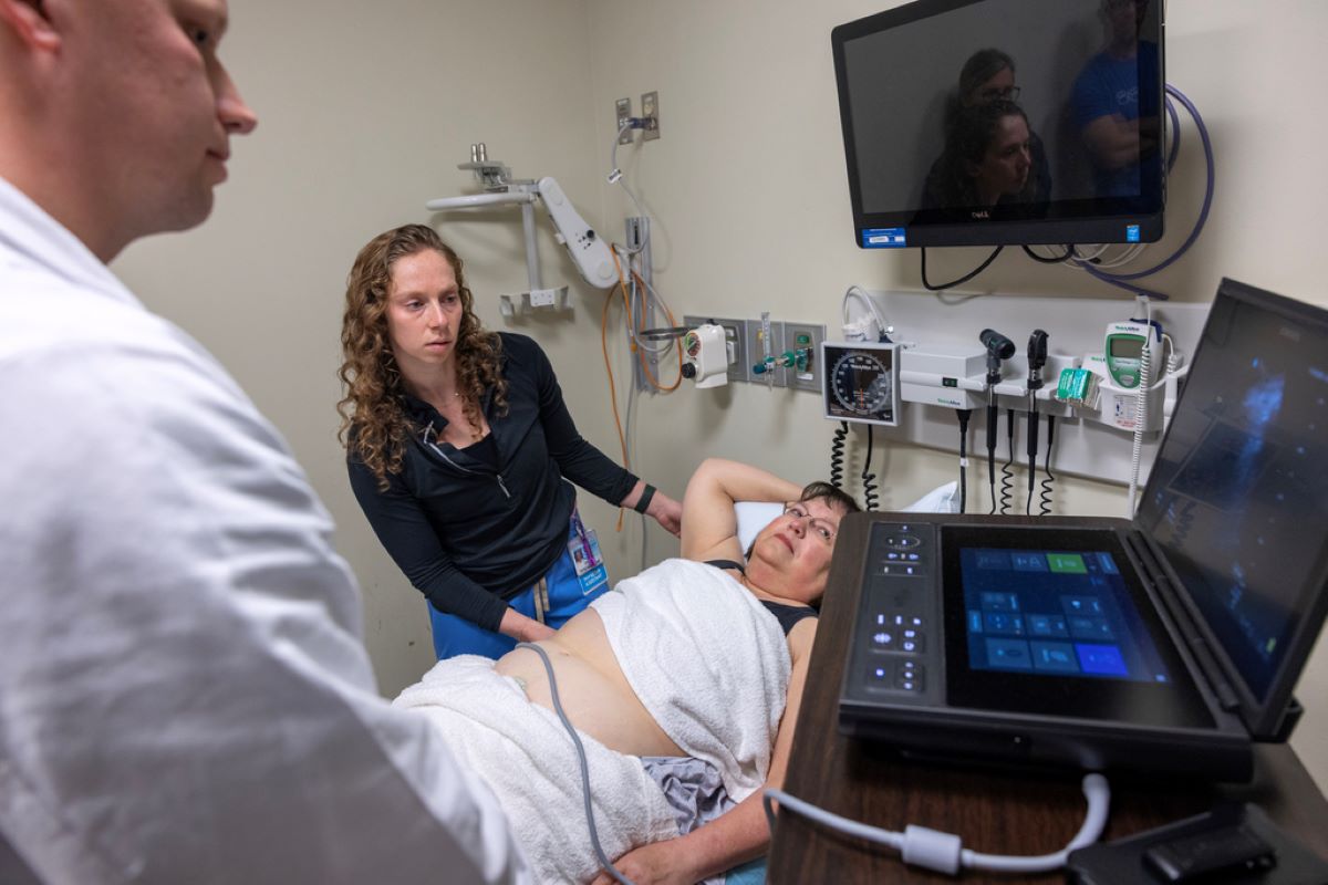 A physician assistant practices an ultrasound on a patient laying in a bed. A man in a white coat stands to the side looking at a medical device screen.