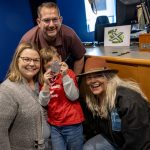 Three adults and one child pose for a photo in a radio studio, smiling.