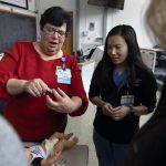 A woman in a red sweater holds a feeding tube and teaches a group of surgical residents.