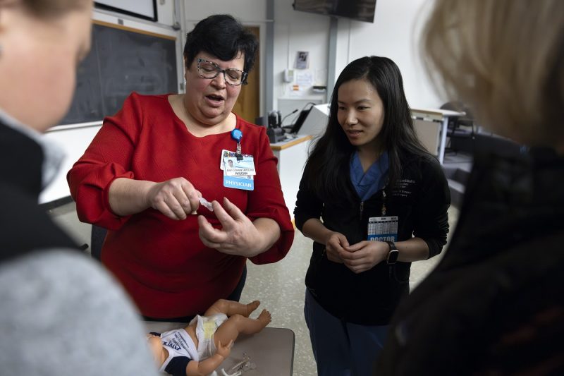 A woman in a red sweater holds a feeding tube and teaches a group of surgical residents.