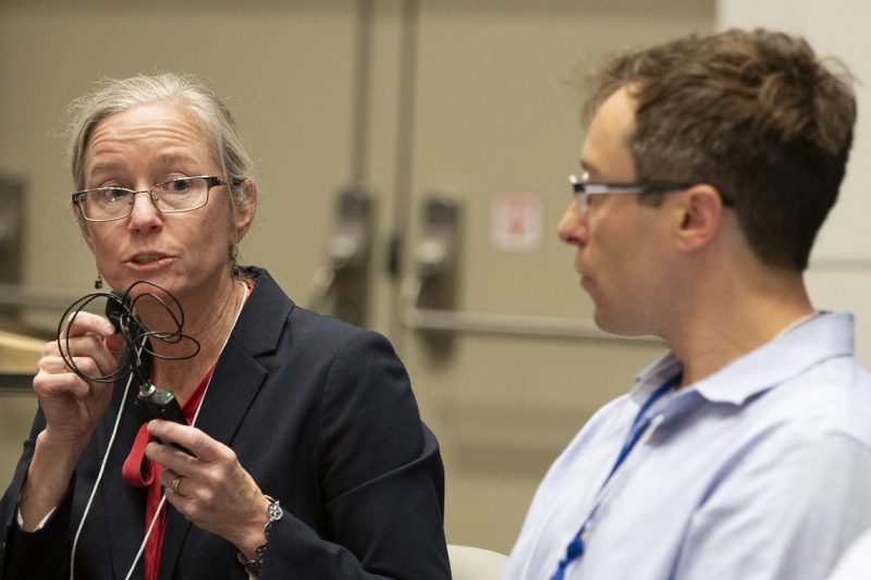 A woman holds a microphone to her face while she speaks. She is sitting next to a man.