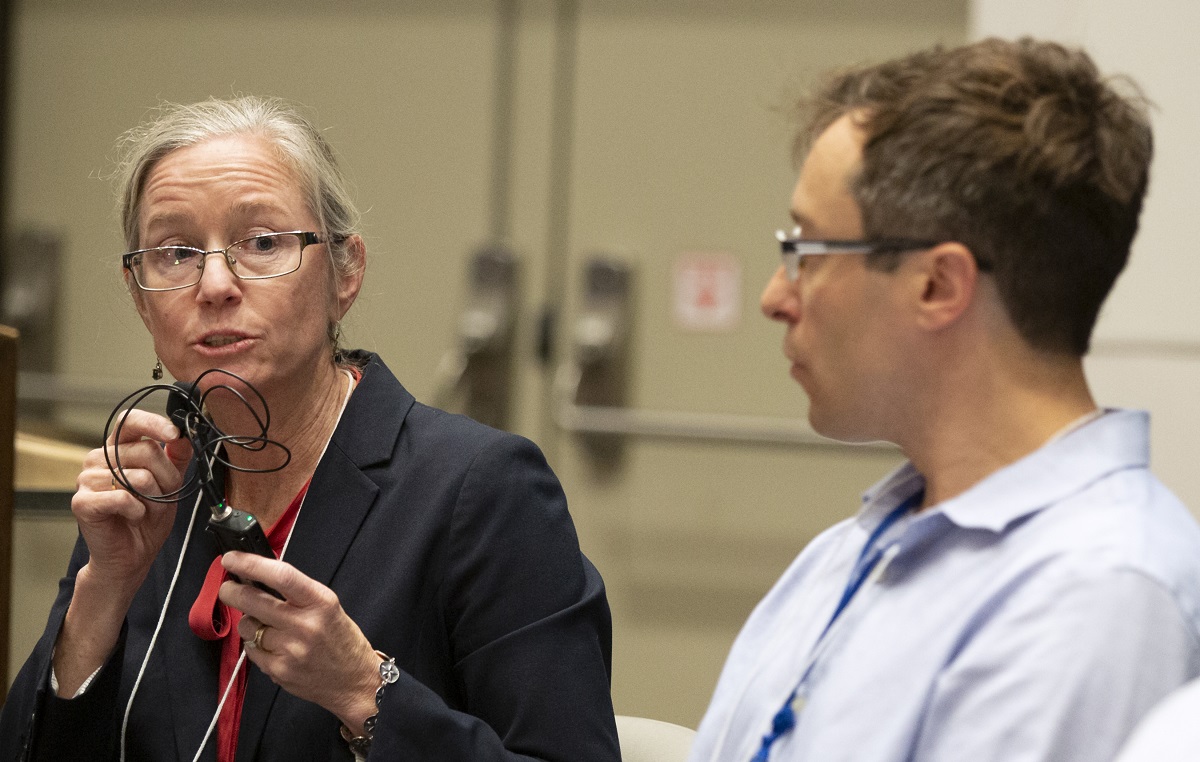 A woman holds a microphone to her face while she speaks. She is sitting next to a man.