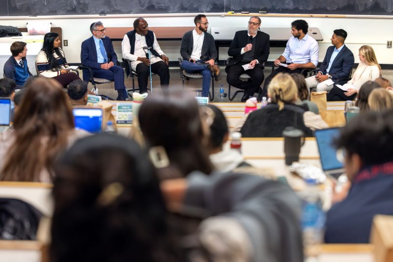 A group of nine individuals is seated in a semi-circle at the front of a large lecture hall.