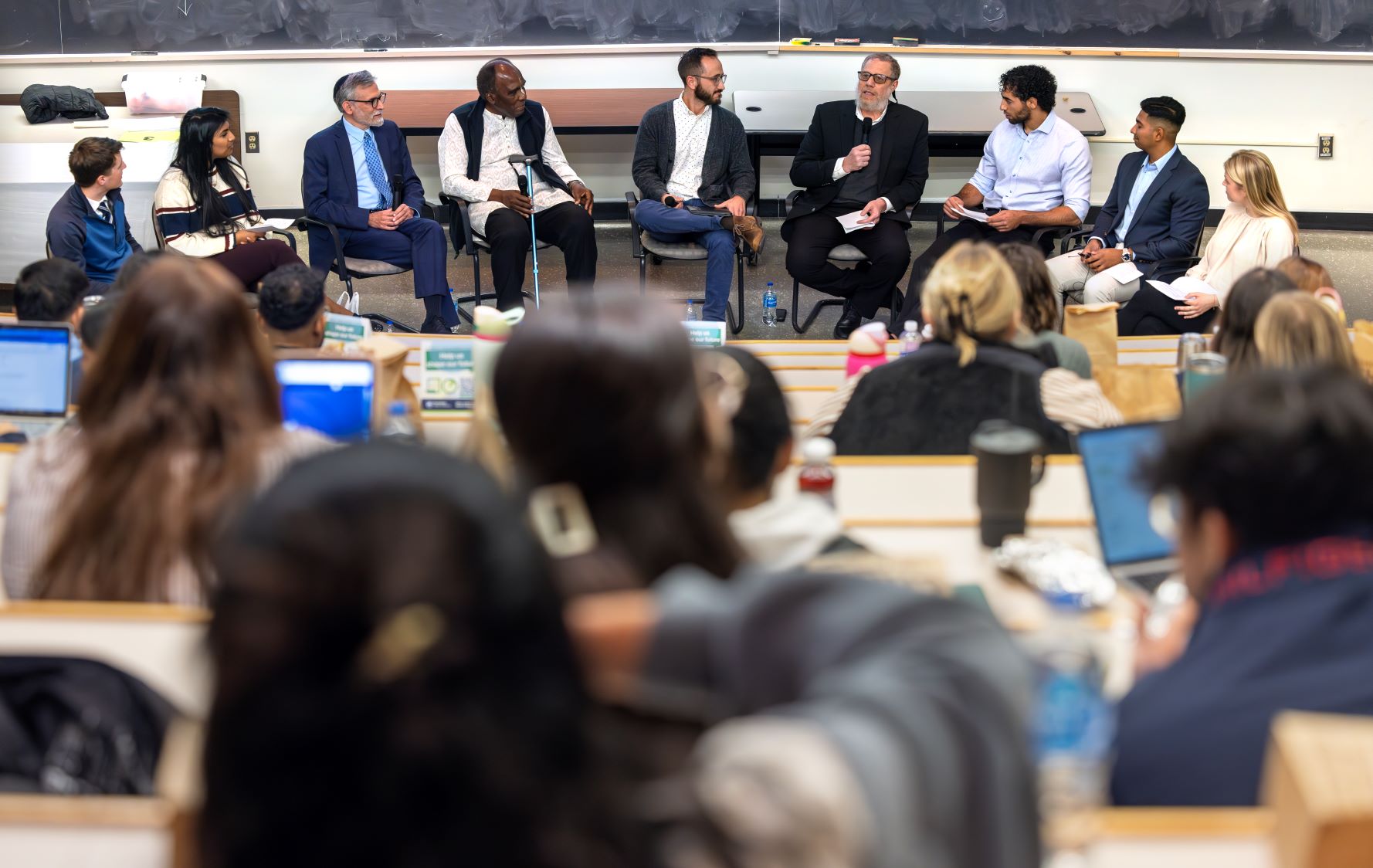 A group of nine individuals is seated in a semi-circle at the front of a large lecture hall.