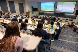 A large lecture hall filled with seated people is seen from the top back row. A group of nine individuals can be seen at the front of the room below large projector screens.