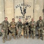 Six U.S. military personnel in camouflage stand together outside, wearing tactical gear, in front of a wall with a “Jackal Ops” logo and “Iraq.”