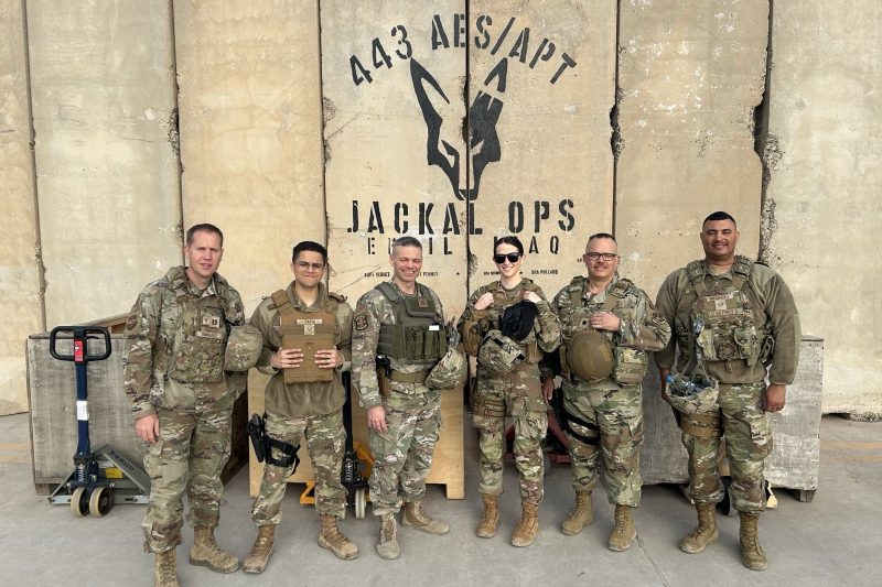 Six U.S. military personnel in camouflage stand together outside, wearing tactical gear, in front of a wall with a “Jackal Ops” logo and “Iraq.”