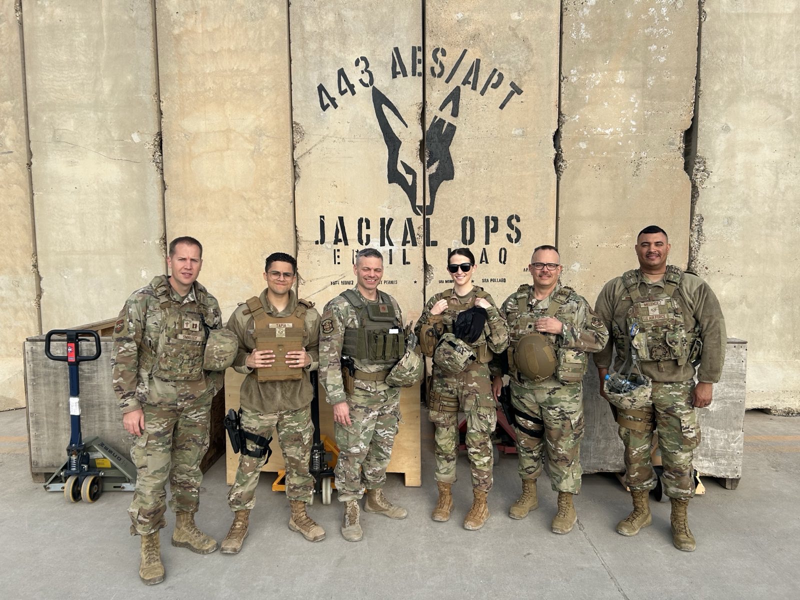 Six U.S. military personnel in camouflage stand together outside, wearing tactical gear, in front of a wall with a “Jackal Ops” logo and “Iraq.”