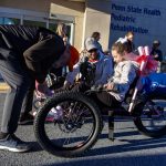 A man leans over to adjust the front of an adaptive bicycle. A young girl sits on the bike smiling, as her dad looks on. Additional people stand in the background.