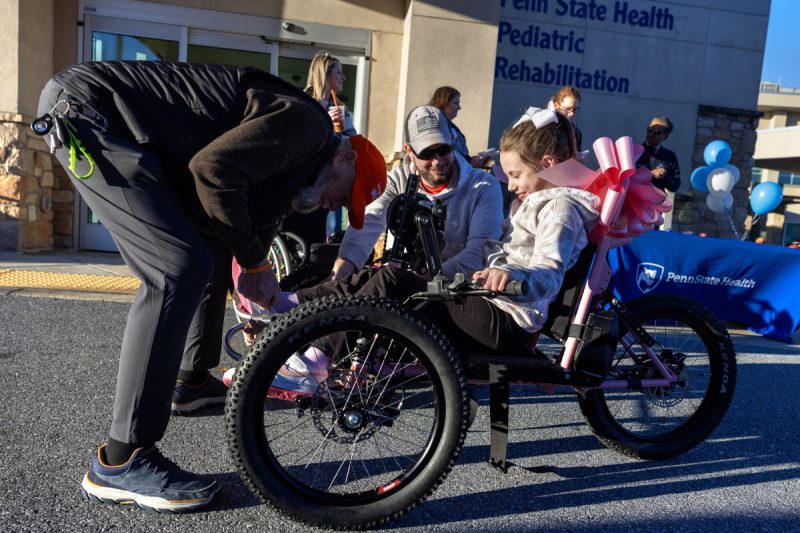 A man leans over to adjust the front of an adaptive bicycle. A young girl sits on the bike smiling, as her dad looks on. Additional people stand in the background.