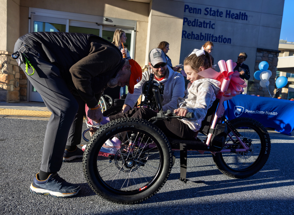 A man leans over to adjust the front of an adaptive bicycle. A young girl sits on the bike smiling, as her dad looks on. Additional people stand in the background.