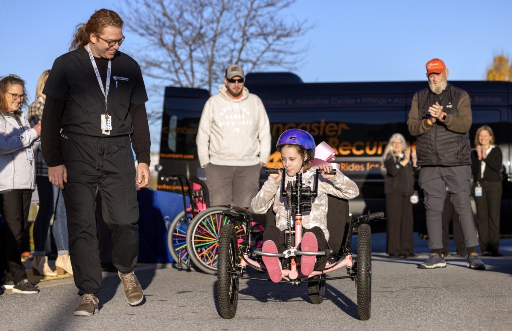 A young girl uses her arms to propel an adaptive three-wheel bike forward. A man in medical scrubs walks alongside the bike as others look on in the background.