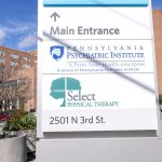 A brick, six-floor hospital is in the background. A sign in the foreground includes the logos for Pennsylvania Psychiatric Institute and Select Physical Therapy.