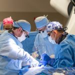 Two doctors perform a transcarotid artery revascularization procedure at Penn State Health St. Joseph Medical Center. They are focused and working in a well-lit operating room, using medical instruments and equipment to carry out the procedure.