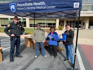 Four people, one in a police uniform, stand under a Penn State Health Hershey Medical Center-branded canopy.