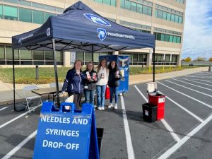 Four people stand under a Penn State Health-branded canopy. A tent sign that says, “Needles and Syringes Drop-Off” sets in the foreground.