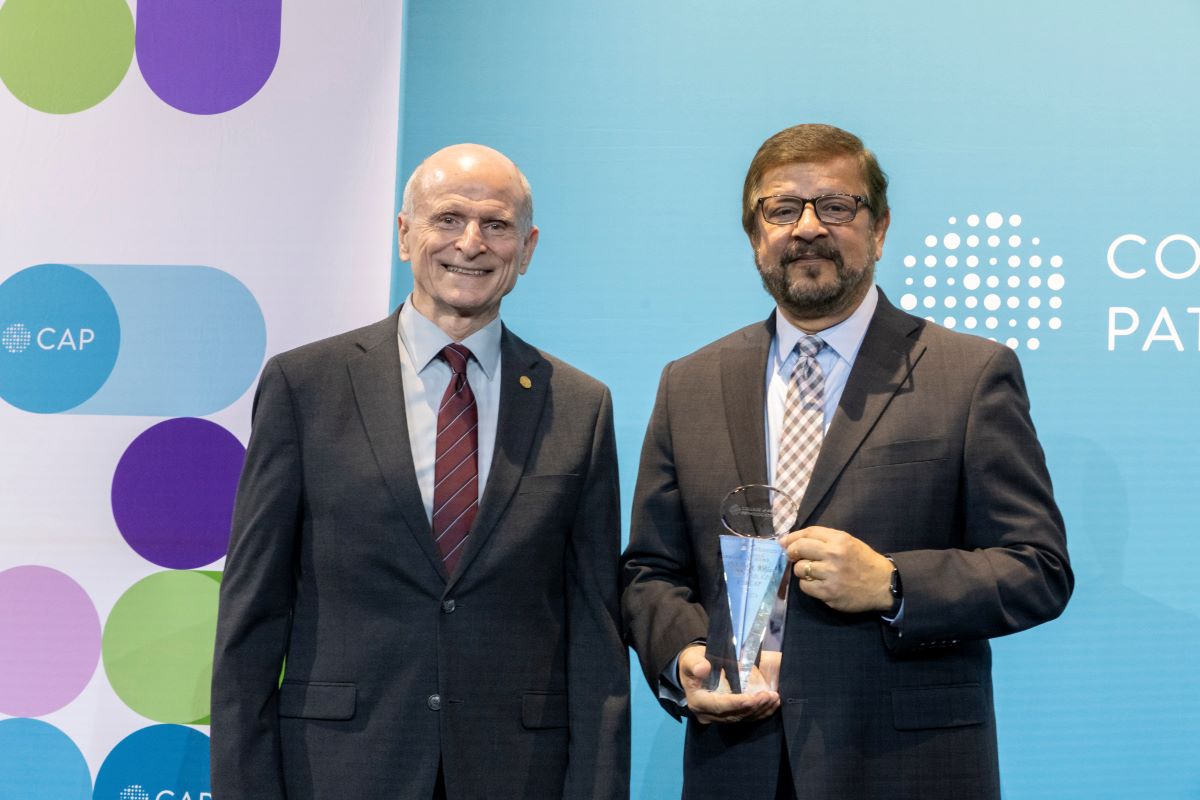 Two men stand side by side. The man on the right is wearing glasses and holding an award. They are both dressed in suits and ties.