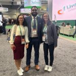 Dr. Nataliya Smith, Dr. Theja Channapragada and Breianna Hummer-Bair stand in the center of a conference center surrounded by exhibits.