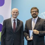 Two men stand on a stage and pose for a photo. The man on the right is holding an award.