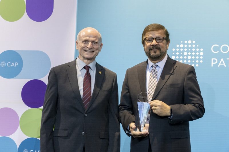 Two men stand on a stage and pose for a photo. The man on the right is holding an award.