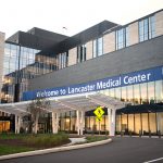 Front entrance of Penn State Health Lancaster Medical Center. The building features a sleek design with extensive glass windows, beige and dark gray brickwork, and a covered entrance with white, curved support beams. A pedestrian crossing sign is visible near the driveway, surrounded by green landscaping and trees.