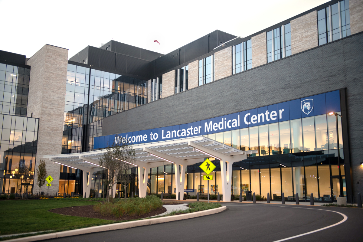 Front entrance of Penn State Health Lancaster Medical Center. The building features a sleek design with extensive glass windows, beige and dark gray brickwork, and a covered entrance with white, curved support beams. A pedestrian crossing sign is visible near the driveway, surrounded by green landscaping and trees.