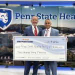 Two men in suits pose with a check made out to “Penn State Health Urology” in the amount of $340,000. A large Penn State Health logo is in the background.