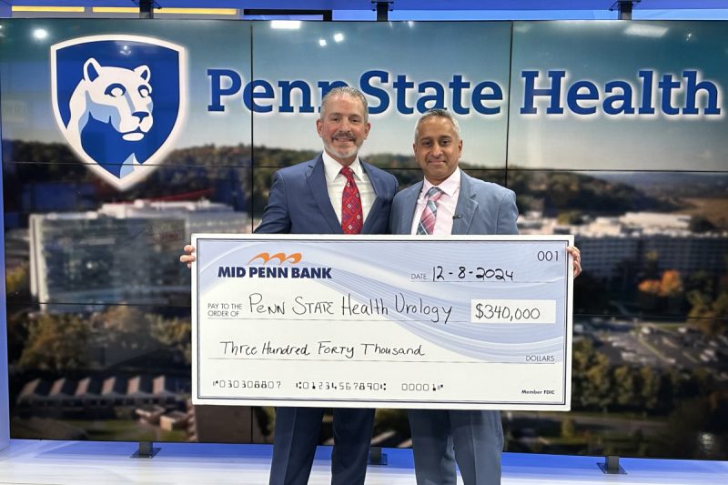 Two men in suits pose with a check made out to “Penn State Health Urology” in the amount of $340,000. A large Penn State Health logo is in the background.