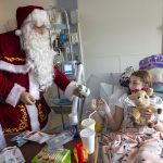 A young girl in a hospital bed, in a room filled with medical equipment and other supplies, smiles as she reached out to accept a gift of a small stuffed animal from Santa Claus.